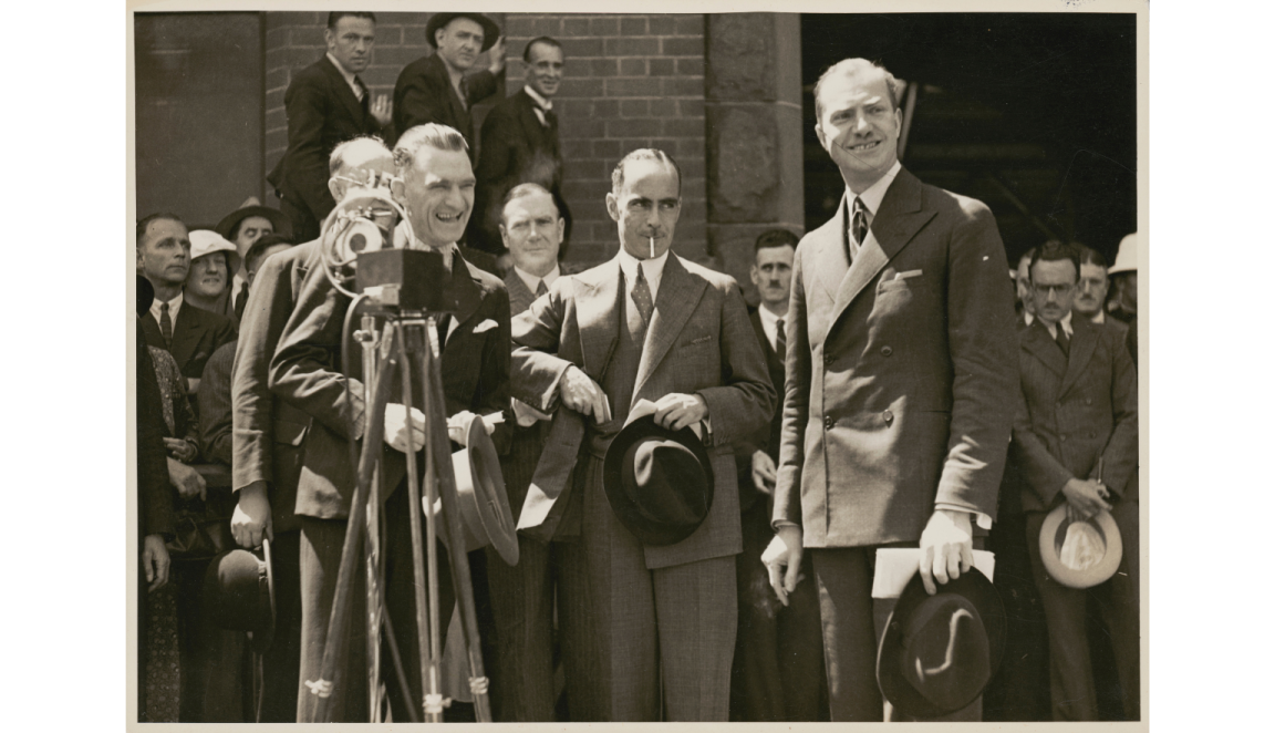 Black and white photo of three men in suits standing in front of a crowd