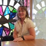 Summer Fellow Deborah Lee Talbot sits at a table in front of the Leonard French stain glass window at the Library. Deborah wears glasses and a white shirt