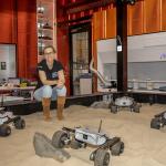 Dr Sarah Cannard sitting on the edge of a sand pit with four space rovers in it