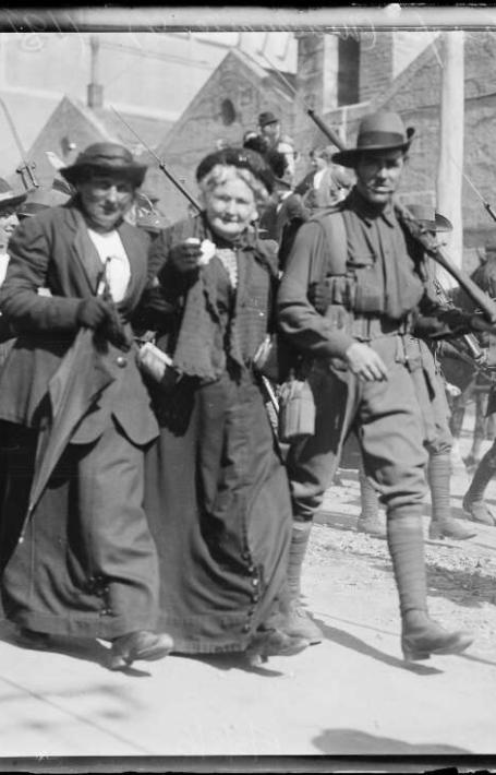 A black-and-white photograph shows a group of soldiers marching down a street with rifles over their shoulders. They are accompanied by civilians, who walk arm-in-arm with the soldiers, smiling and engaging with the crowd. The street is busy with people, and buildings can be seen in the background.