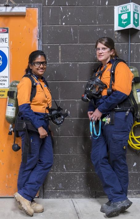 Two women in navy and orange work wear with protective glasses, gas masks and tanks on their backs standing by a wall with an orange door and signs reading 'Danger Chlorine', 'Eye protection must be worn in this area', 'Liquid chlorine bulk storage' and more.