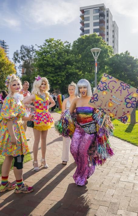 A group of four people who are very colourfully dressed, holding signs in a park. There are more groups of colourful people in the background.