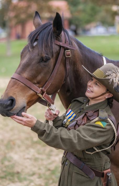 Female solider in uniform smiling and patting a military horse, with another soldier in uniform holding the reins of another horse nearby