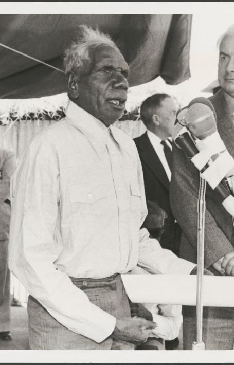 Vincent Lingiari addresses a crowd at a microphone while Gough Whitlam stands beside him. Several people are visible in the background, observing.