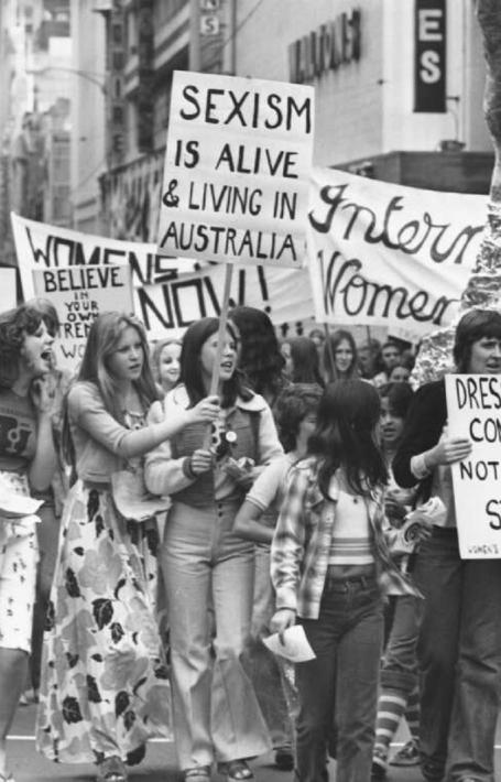 A group of women marches down a city street, holding various protest signs. Signs include messages like "Sexism is alive & living in Australia" and "Dress for comfort, not for style." A large sculpture is visible in the background.