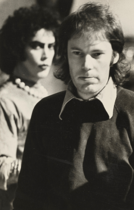 Black and white photo of Jim Sharman walking towards the camera while an actor dressed as Dr Frank-N-Furter in The Rocky Horror Picture Show stands behind him