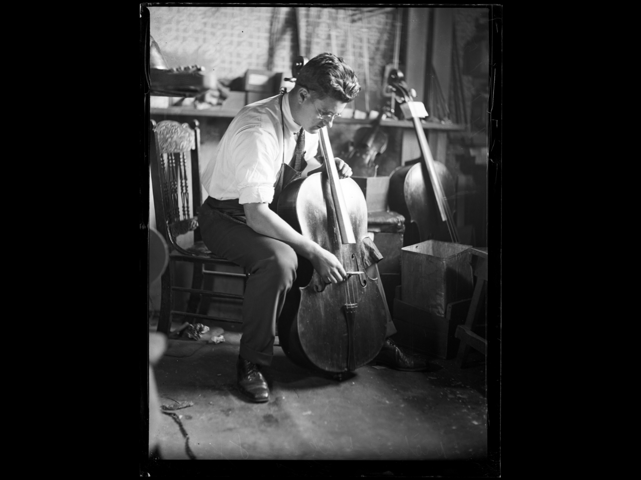 Black and white photo of a man sitting and examining a cello in a workshop