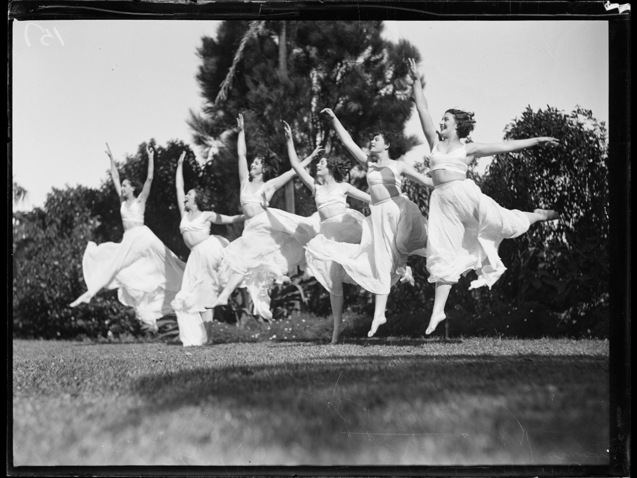 Black and white photo of six dance students in white tops and skirts leaping