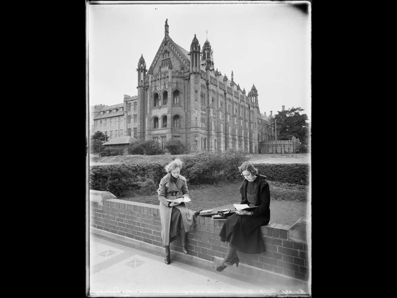 Black and white photo of two young women sitting and reading on a wall in front of a large ornate building