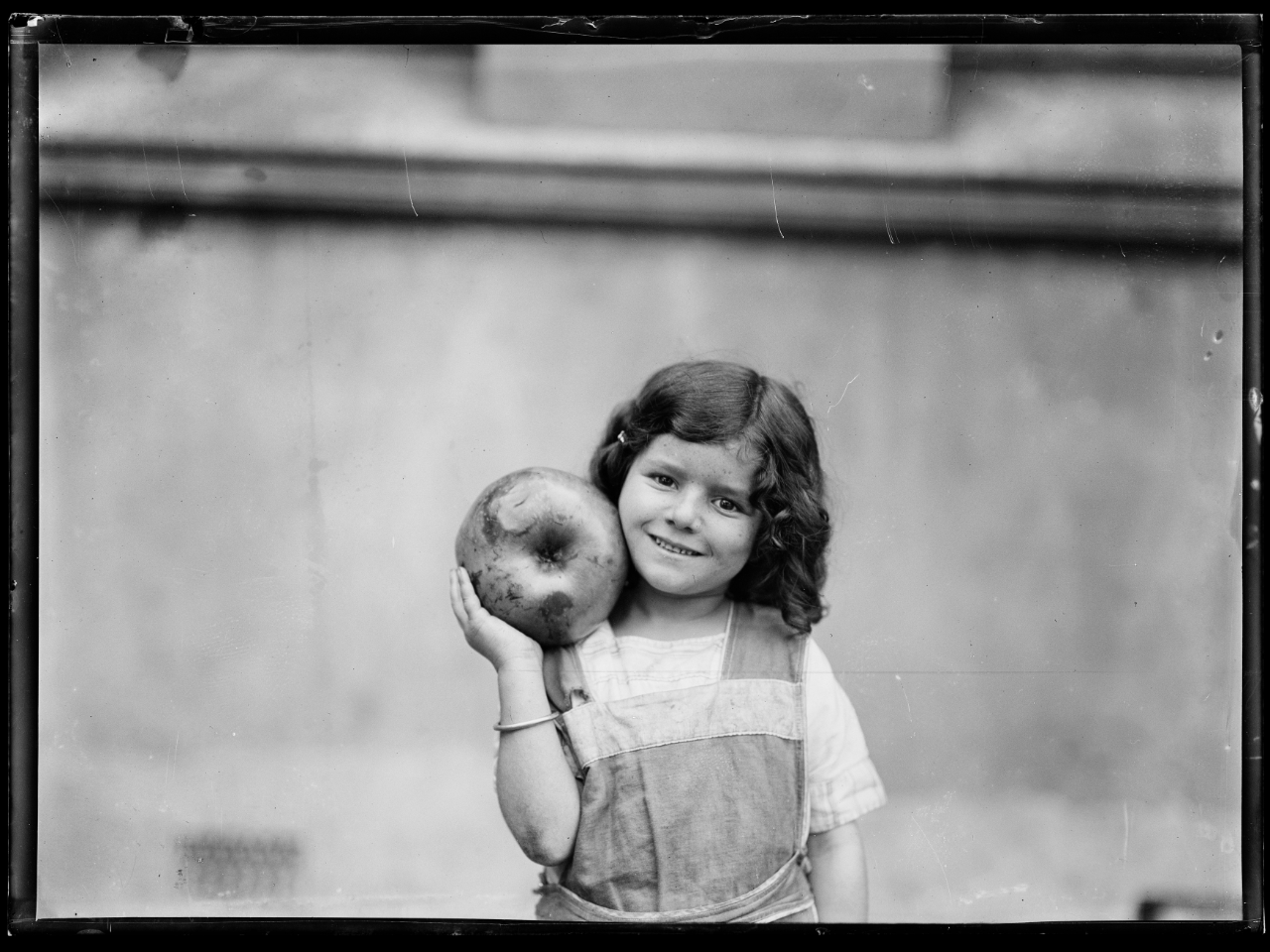 Black and white image of a young girl smilng and holding a large apple up on her shoulder
