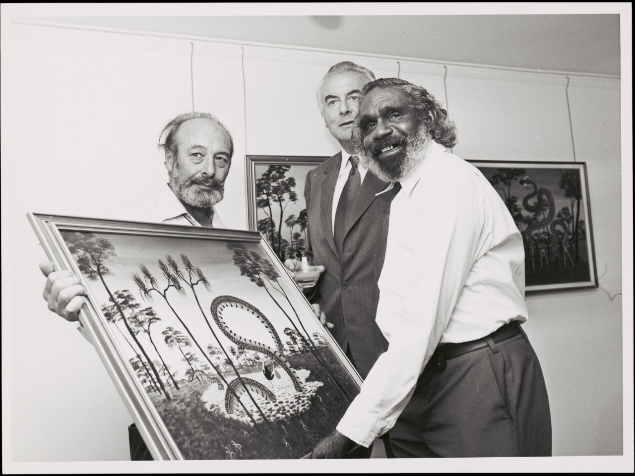Black and white photo of Percy Trezise and Dick Roughsey holding a large painting of the Rainbow Serpent next to then Prime Minister Gough Whitlam