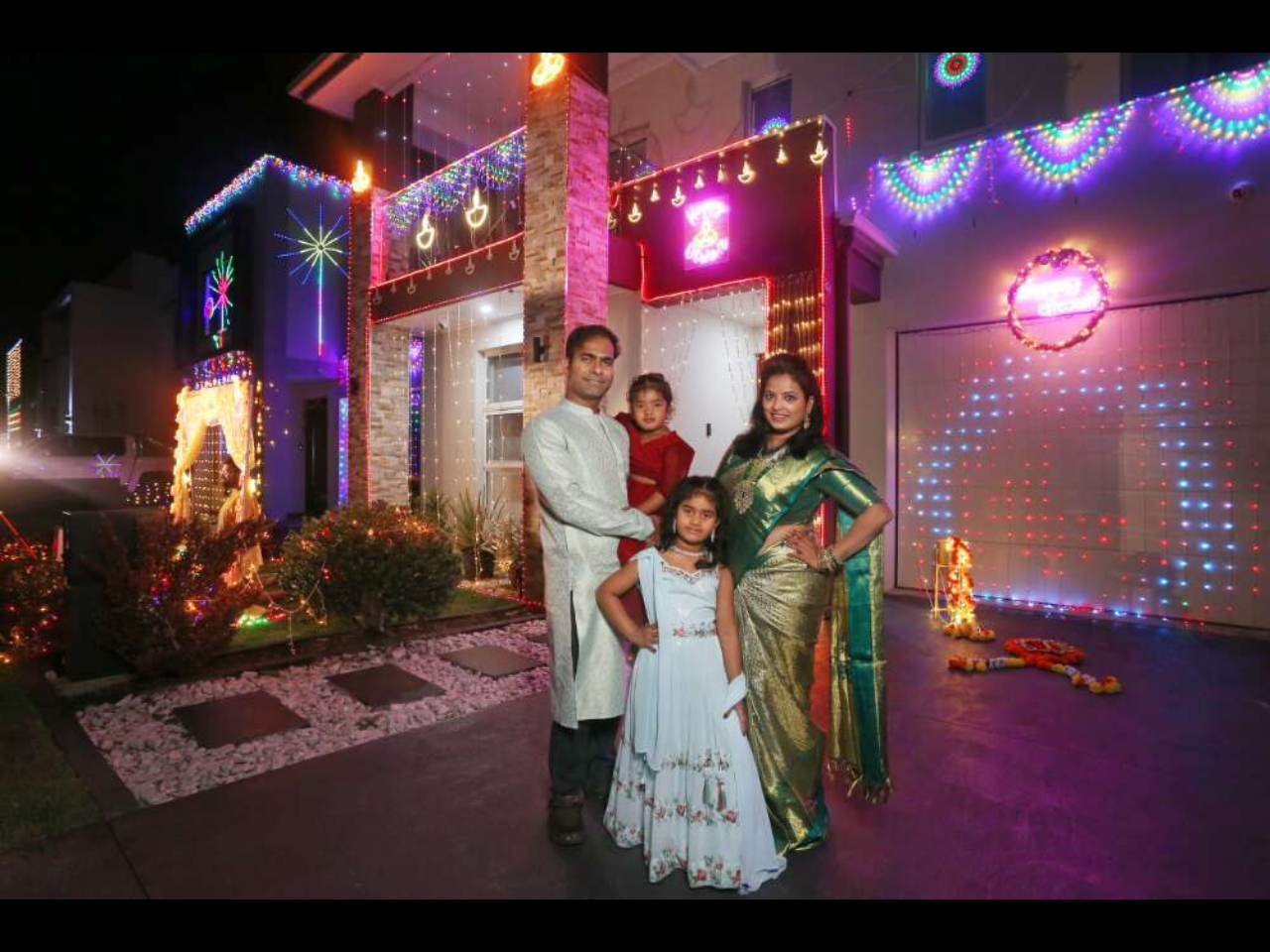 A family of four standing in front of their house. Their house is decorated with coloured lights.