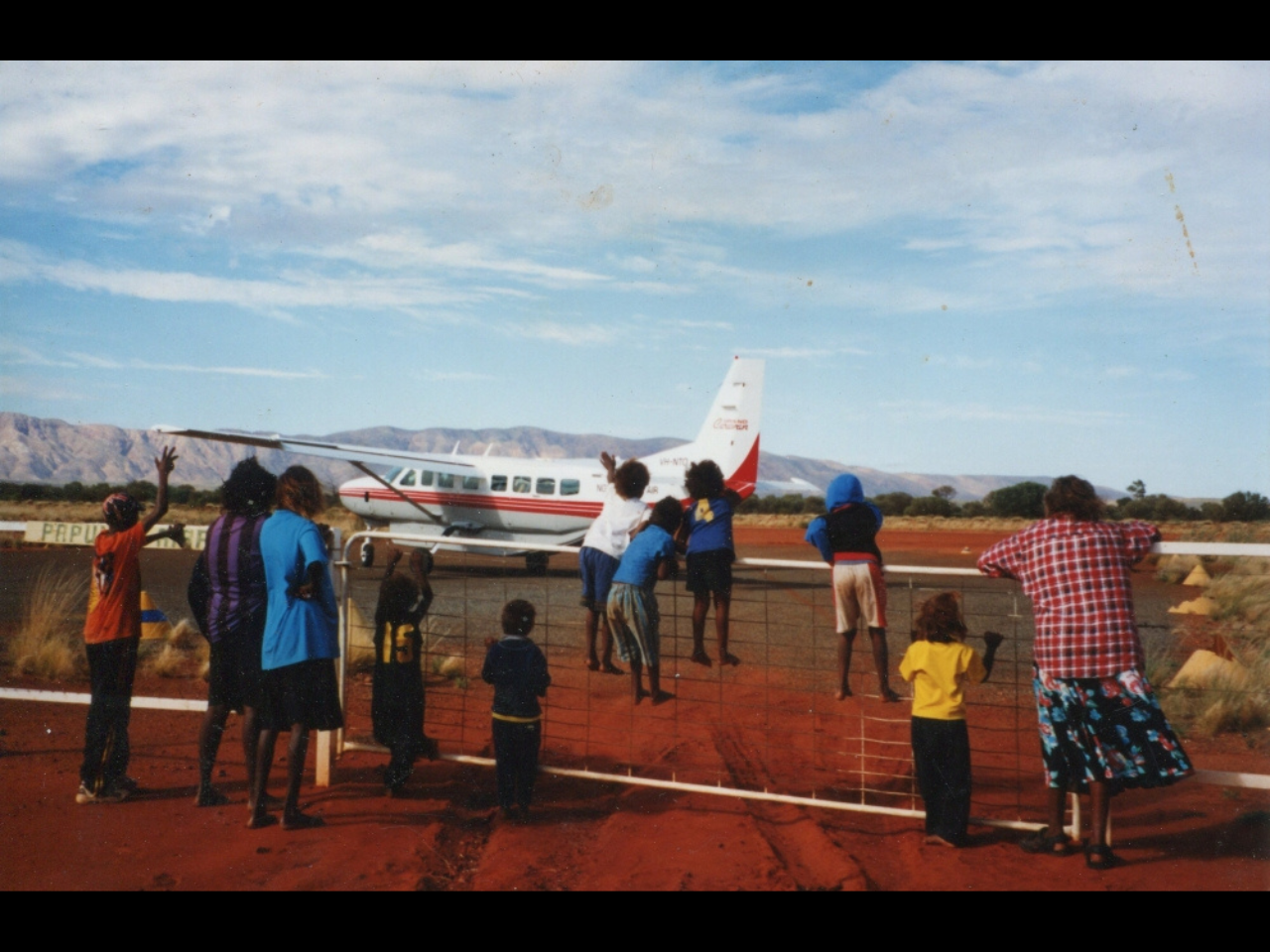 Several people of varying age leaning on a fence looking at a plan on an airstrip