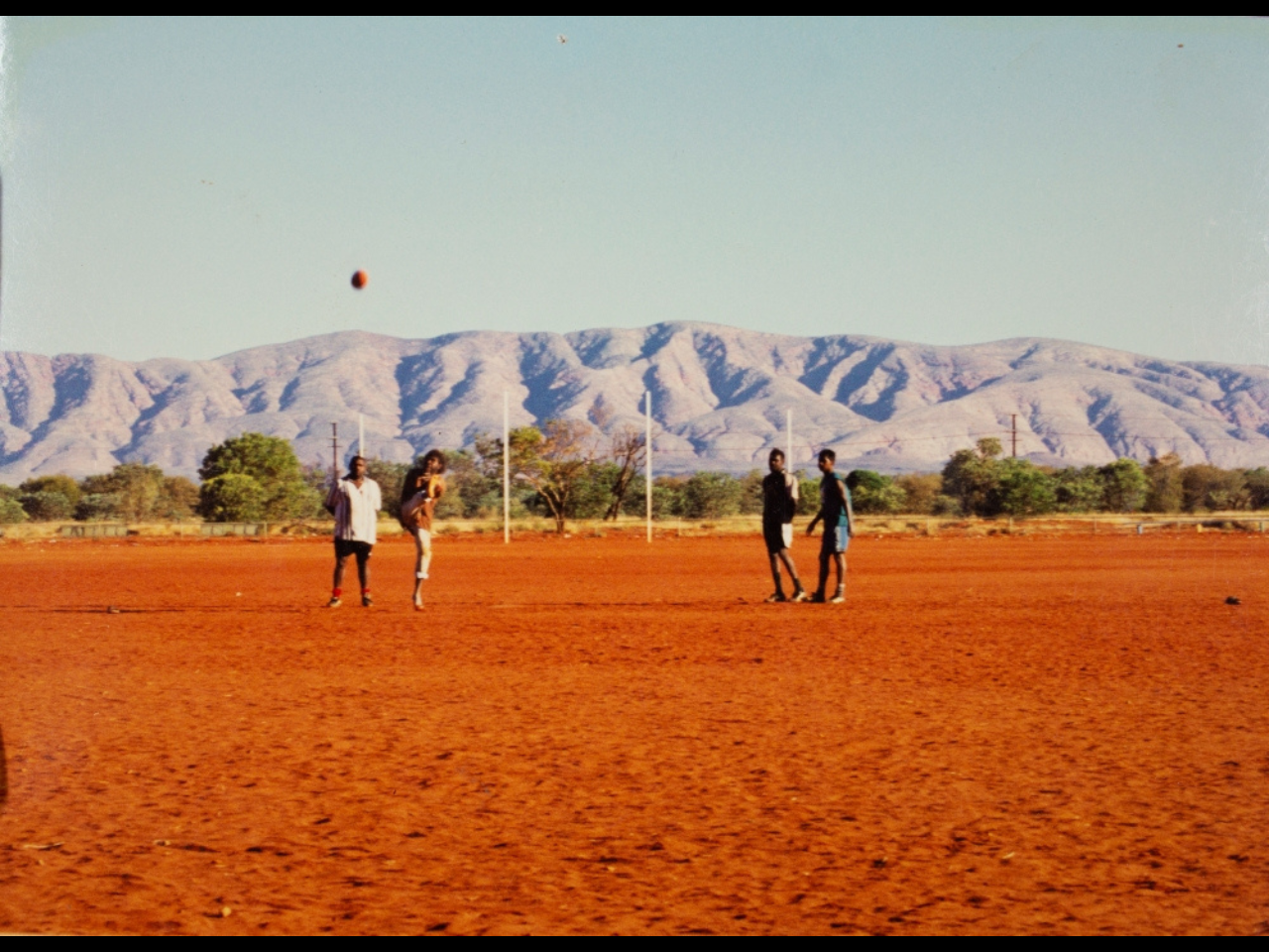 Four people play AFL football on a field. There are hills in the background.