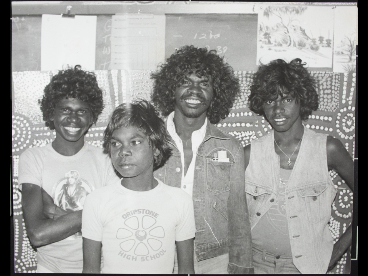 Four people stand in front of a wall in this photo. There is artwork, a chalkboard and papers attached to the wall behind them.
