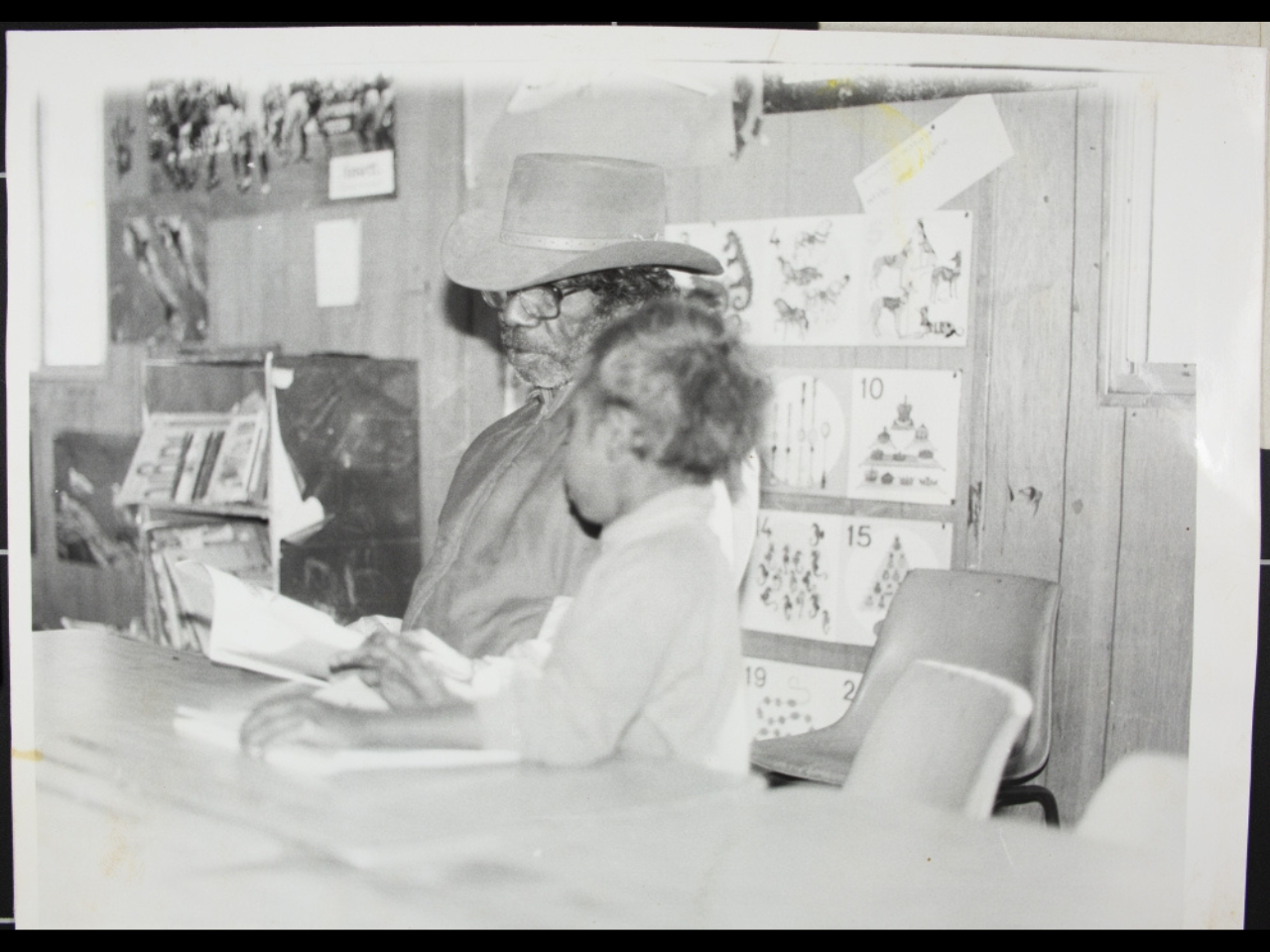A man and a child sit at a desk in a classroom reading a book.