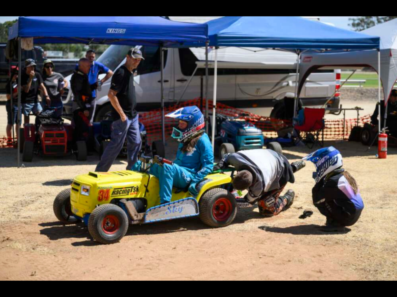 A young person sits on a ride on lawn mower while repairs are conducted on it. A group of other people look on.