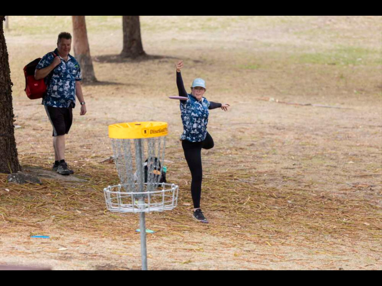 A disc golfer stands on one leg, their disc mid-flight towards the basket. A second golfer standing next to a tree behind them watches on.