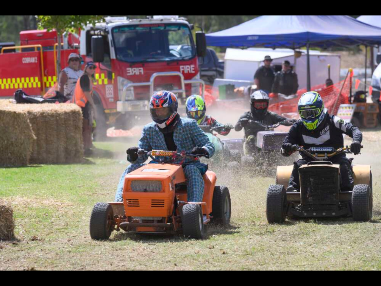 Four individuals race ride-on lawn mowers on a grassy field. A fire truck is visible in the background.