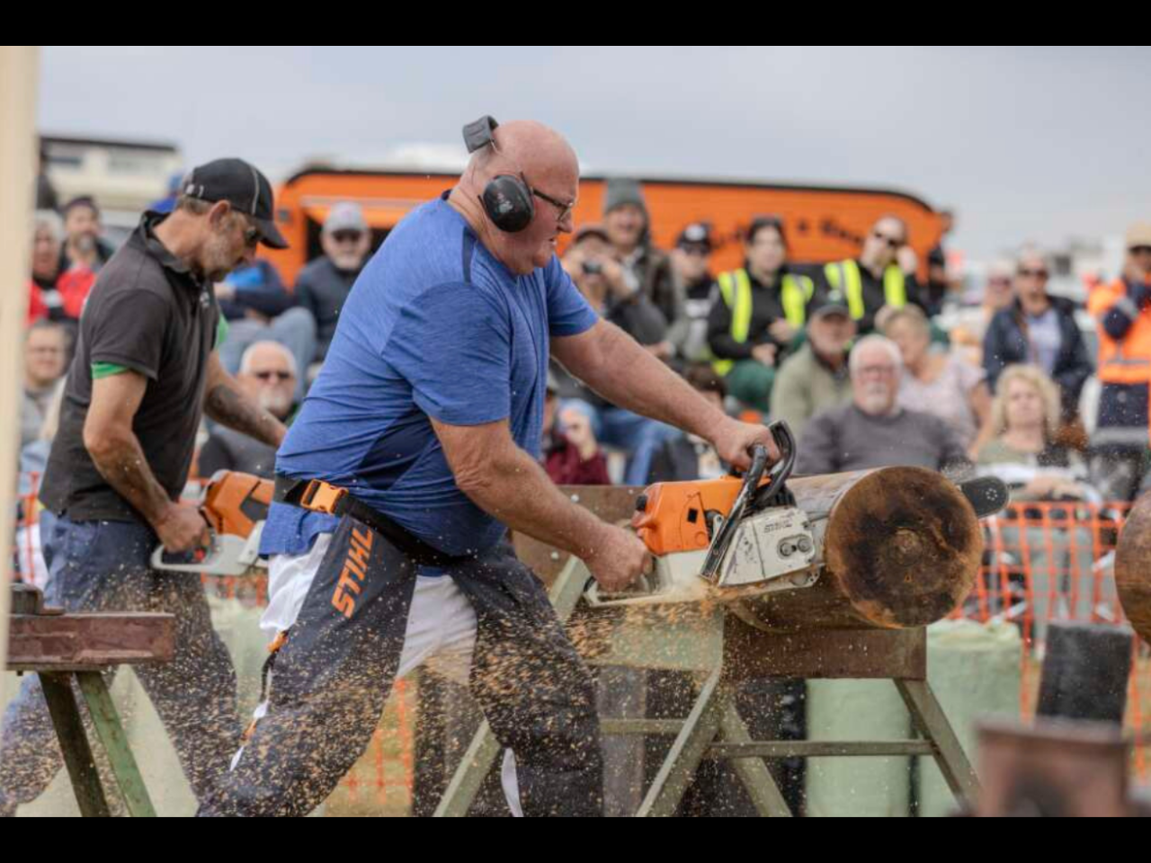 A man is using a chainsaw to cut through a piece of wood, generating a cloud of sawdust. Several other people are also using saws. A crowd watches in the background.