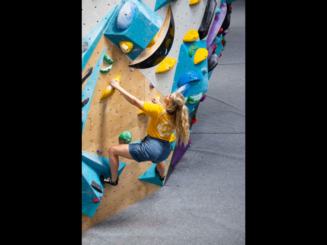 A woman in a yellow shirt and denim shorts is climbing an indoor bouldering wall. 