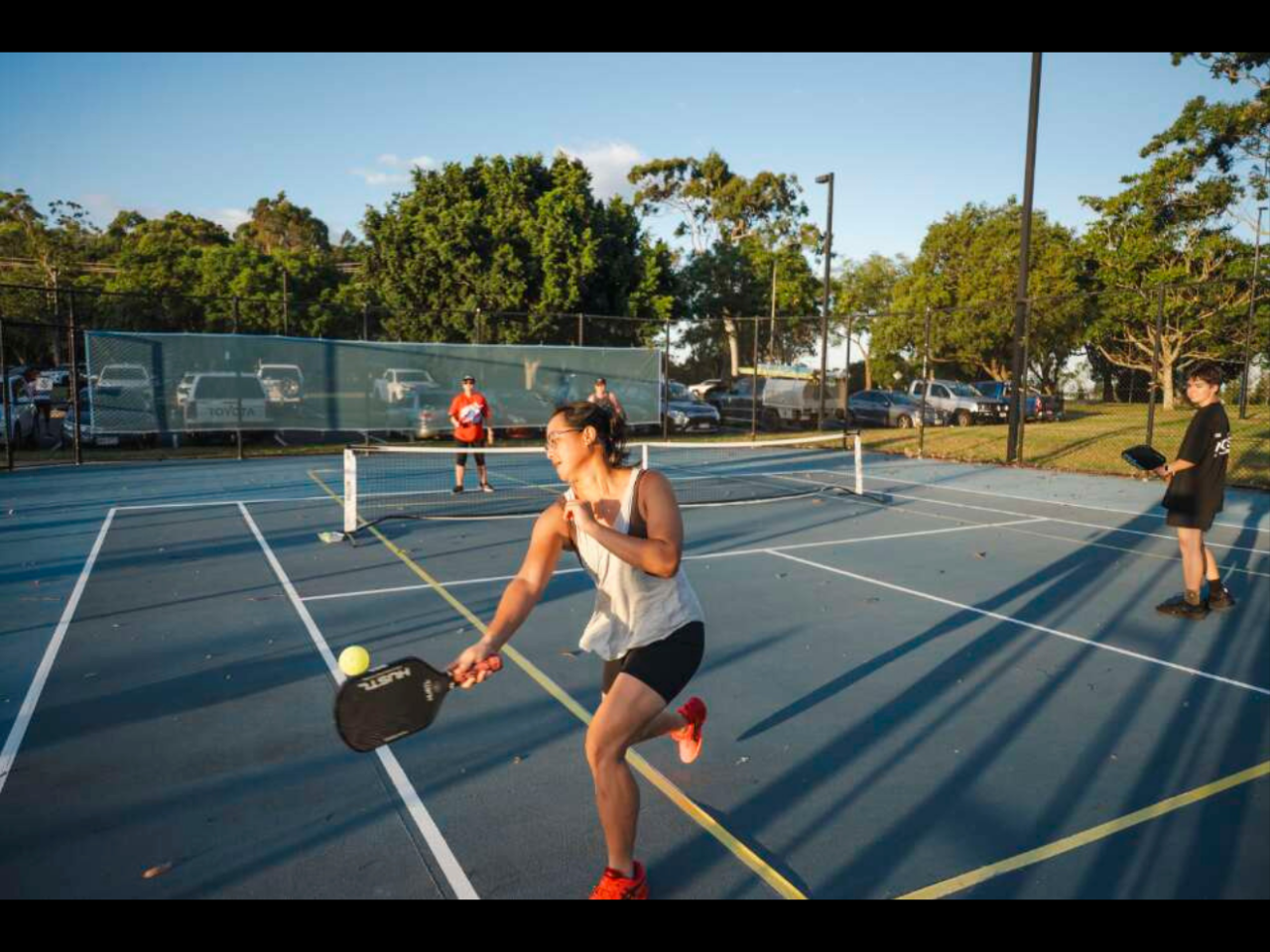 A woman is playing pickleball on an outdoor court. She is mid-swing, focused on hitting the ball, while other players stand nearby. The court is surrounded by a fence with trees in the background.