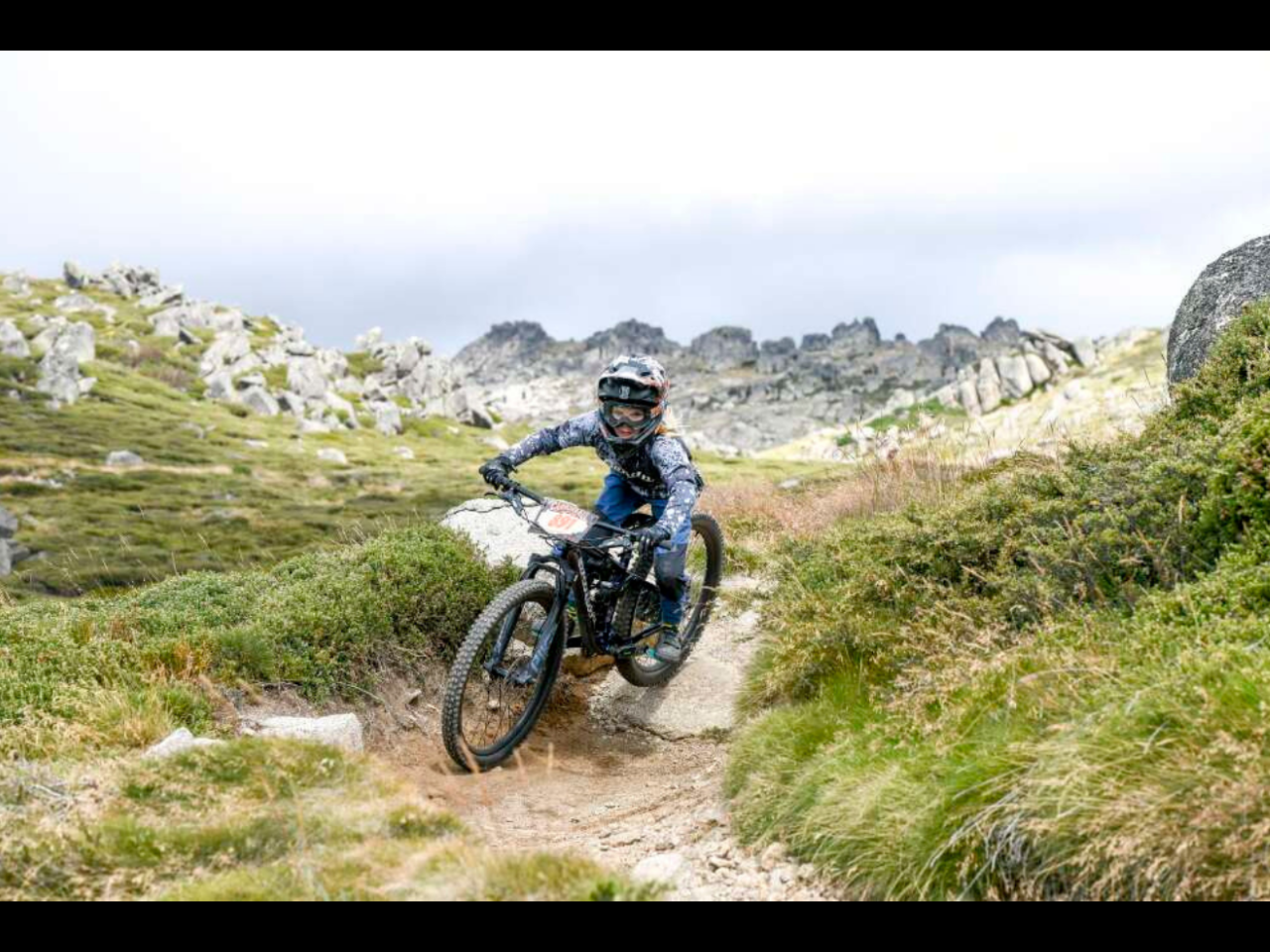 A mountain biker wearing full protective gear rides a bike down a rugged, grassy trail under an overcast sky. The landscape features rolling hills and rocky outcrops.