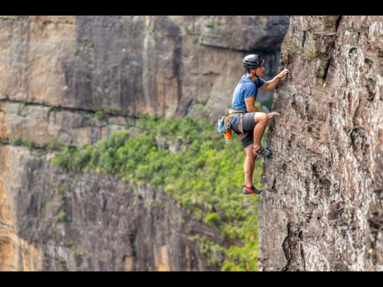 A rock climber is scaling a large, textured rock face outdoors. Cliffs featuring green foliage are visible in the background. 