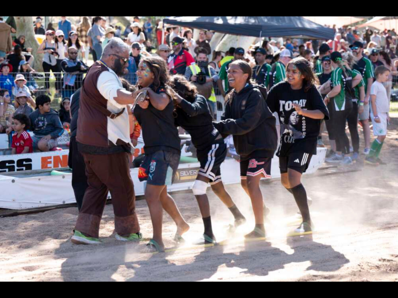 A group of young people laugh as they participate in an outdoor race on a dusty riverbed. 
