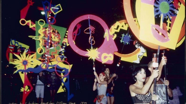 Participants in the 1995 Sydney Gay and Lesbian Mardi Gras parade carry large, colourful, abstract posters, flags and sculptures. The scene is set at night with vibrant shapes, patterns, and faces on display.