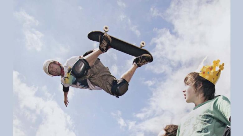 A person on a skateboard is in the air. Two people in the bottom right hand corner are looking up at him. One of them is wearing a cardboard crown. The background is a blue sky with white clouds.
