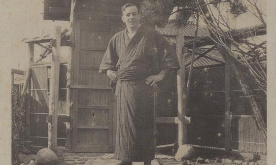 A sepia photograph of a man in traditional Japanese dress standing outside in front of a wooden dwelling structure.