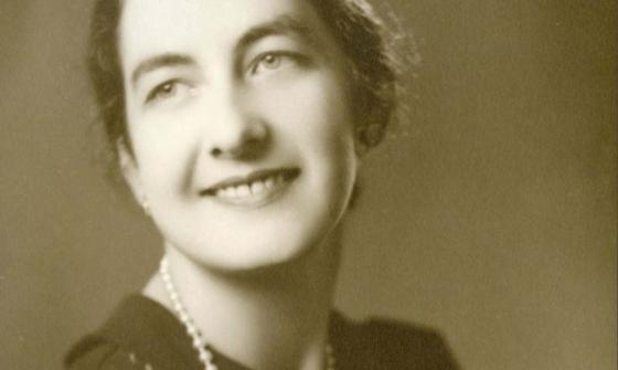 A sepia image of a woman in a black dress and pearl necklace, looking back and smiling over her shoulder.