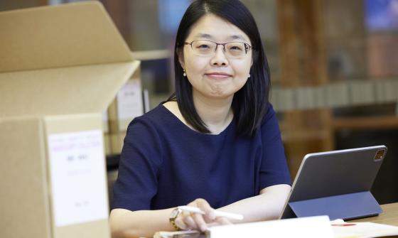 Woman with short dark hair wearing a navy short sleeve top smiling at the camera and sitting at a desk with a laptop, papers and a box