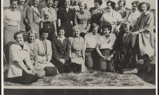 Black and white photo of 21 women of various ages smiling, standing in front of a plane. Below the image is a list of these women's names and where they're from
