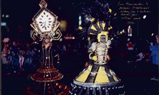 Two people in a night time parade dressed up in large, intricate, colourful costumes.