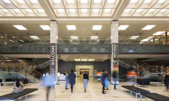 Large two-story room with cream-coloured marble floors, columns and stairs; black leather couches; lots of light from the ceiling and a balcony from the second floor looking over the room. On the columns are signs with directions and the word 'Welcome'. People are moving around the room.