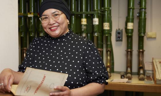 Woman wearing a dot print shirt, glasses and a black head scarf holding an open booklet and smiling