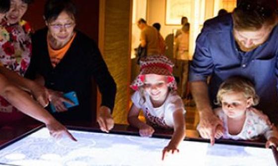 A group of people, including two children and three adults, engaging with an interactive exhibit. They are gathered around a glowing display table, pointing and exploring the content.