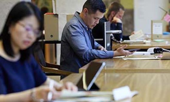 A group of people working in the special collections reading room. One person in the foreground is writing in a notebook, another in the middle is using a smartphone while taking notes, and others in the background are focused on their work.