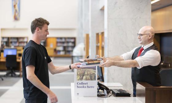 A person in a black T-shirt hands over several books to a staff member wearing a white shirt, black vest and red tie. They stand at a library desk labelled "Collect books here" with a computer and phone visible on the counter.
