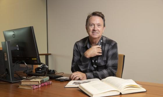 A man wearing a black and grey checkered shirt sitting at a desk with a computer and several books resting on it. He is leaning on the desk, facing forward and giving the camera a small smile
