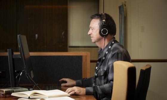 Man in a grey and black checkered shirt and headphones sitting at a desk, using a computer. To his left are two open books