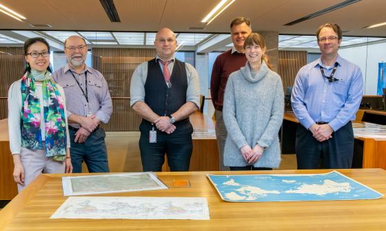 Two women and four men, who make up the Library's Special Collections Research & Support team, standing and smiling in the Special Collections Reading Room in front of a table with three maps on it