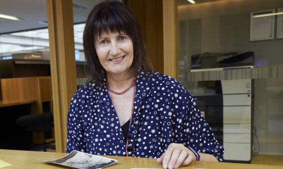 Smiling woman with short black hair and a fringe sitting at a table where several black and white photos of a woman are spread out