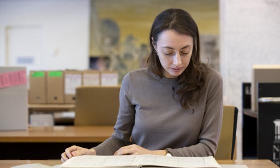 Woman with brown hair and eyes, wearing a brown sweater, sitting at a desk in the Special Collection Reading Room, looking down as she reads from an old book.