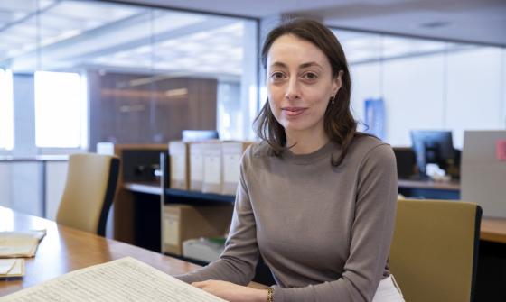 Woman with brown hair and eyes, wearing a brown sweater, sitting at a desk in the Special Collection Reading Room, smiling at the camera. On the desk in front of her is an old book of music.