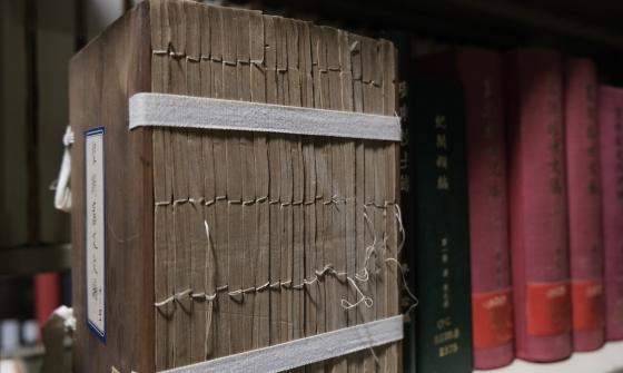 A large, old text with Chinese characters is bound with traditional thread stitching, sitting on a shelf, supported by a white fabric strap wrapped around its cover. The edges of the pages are uneven, showing wear with the stitching slightly frayed. Other colourful books with Chinese characters are lined up alongside it on the shelf.