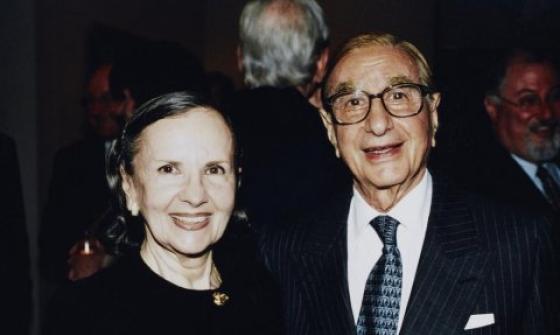 A couple smiles at the camera in a formal setting. Susan Wakil (AO), with dark hair pulled back, wears a black outfit with gold buttons. Isaac Wakil (AO), wearing glasses and a dark pinstriped suit, holds a wine glass in one hand. Both appear happy and well-dressed, surrounded by a dimly lit background with other people.