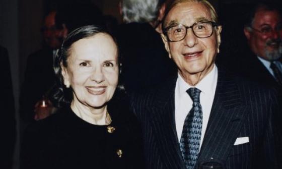 A couple smiles at the camera in a formal setting. Susan, with dark hair pulled back, wears a black outfit with gold buttons. Isaac, wearing glasses and a dark pinstriped suit, holds a wine glass in one hand. They are surrounded by a dimly lit background with other people.